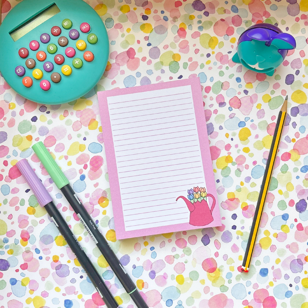 Colourful flat lay with a round teal calculator, purple sharpener, black pencil, two markers, and the Pink Gardening A6 Memo Pad—featuring a pink watering can—arranged on a pastel spotted background.