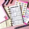 A personal ring planner open on a table. The top sheet shows a 'reading journal' insert. Around the planner are sticker sheets, a pink memo pad and some pens.