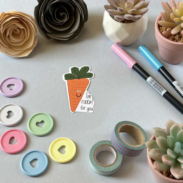 A flat lay of craft supplies features pastel heart-shaped buttons, washi tape rolls, two pens, small potted succulents, paper flowers, and the Rootin For You Matt Vinyl Sticker—a cheerful smiling carrot with "I'm rootin' for you" text.