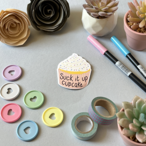 A light grey background with a single sticker lay in the centre. A cupcake with a pink case and a swirl of white frosting on top with some rainbow sprinkles on it. On the case it says 'suck it up cupcake'. Around the edge are paper roses, washi tape, planner discs and brush pens.
