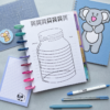 The Savings Tracker Planner Insert 3 with a jar illustration in a colourful binder is on a desk, surrounded by a blue koala notepad, a small sheep note card, pens, and a cutting mat featuring a bear sticker.
