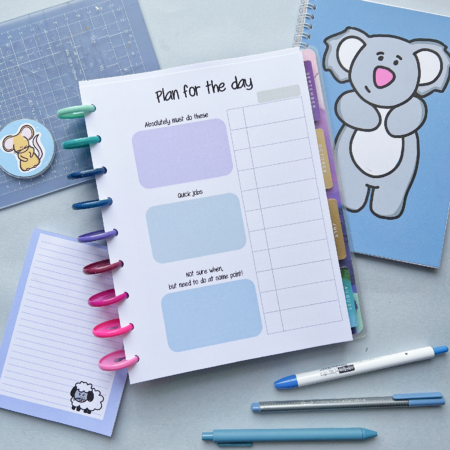 A daily planner with sections for priorities, quick jobs, and other tasks is open on a desk along with pens, stickers, a cutting mat, a koala notebook, a sheep notepad, and the Schedule and Tasks Insert 2.