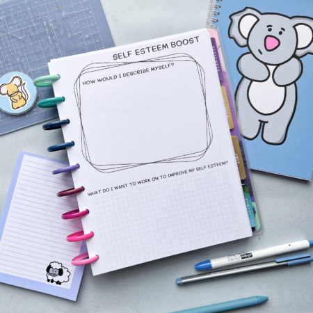 A classic disc planner is open on a table. The top sheet says 'self esteem boost' and then has two box areas. The top says 'how would I describe myself' and has three box outlines that are fanned out over one another. The bottom section says 'what do I want to work on to improve my self esteem?' and has a blank grid area for writing on. Around the planner is various blue stationery including blue pens, a memo pad, a keyring and a cutting mat. 