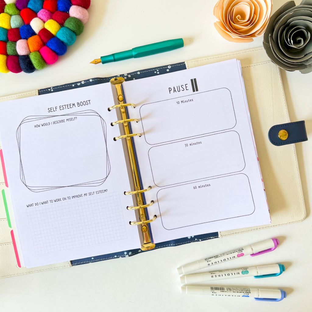 The Self Care Tracker Planner Sheet 3 is open to sections for self-esteem and pause exercises, displayed with colourful felt coasters, a teal pen, two marker pens, and paper flowers on a white surface.