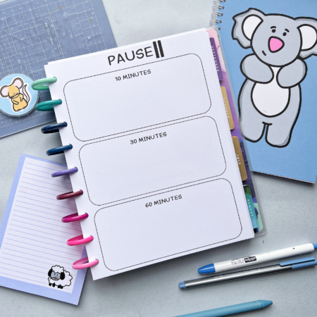 A classic disc planner is open on a table. The top sheet has a title of 'PAUSE' with a pause sign next to it. Below are three outlines of boxes. In each box is a title of 10 minutes, 30 minutes and 60 minutes in the bottom one. Around the planner is various blue stationery including blue pens, a memo pad, a keyring and a cutting mat. 