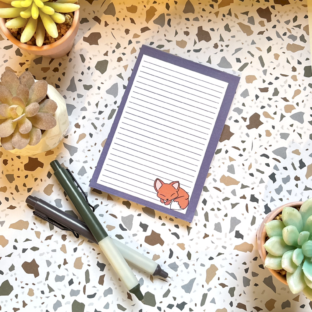 The Sleepy Fox A6 Memo Pad with lined pages rests on a terrazzo surface, accompanied by three potted succulents and two pens—one black, one green.