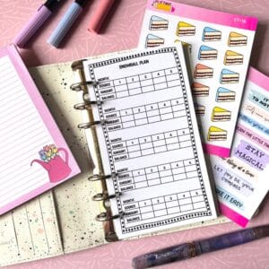 A personal ring planner open on a table. The top sheet shows a 'snowball plan' insert. Around the planner are sticker sheets, a pink memo pad and some pens.