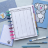 The Spending Tracker 2 binder with a financial ledger sheet and planner insert sits on a desk, surrounded by colourful pens, a blue koala notebook, a sheep notepad, a sticker, and a cutting mat.