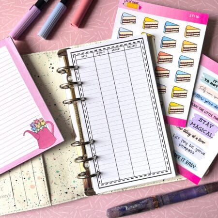A personal ring planner open on a table. The top sheet shows a 'spending tracker' insert. Around the planner are sticker sheets, a pink memo pad and some pens.
