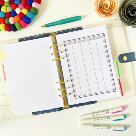 The Spending Tracker is open to a blank sheet and a spending tracker insert, surrounded by three pastel markers, a green pen, colourful felt coaster, and paper flowers on a white surface.