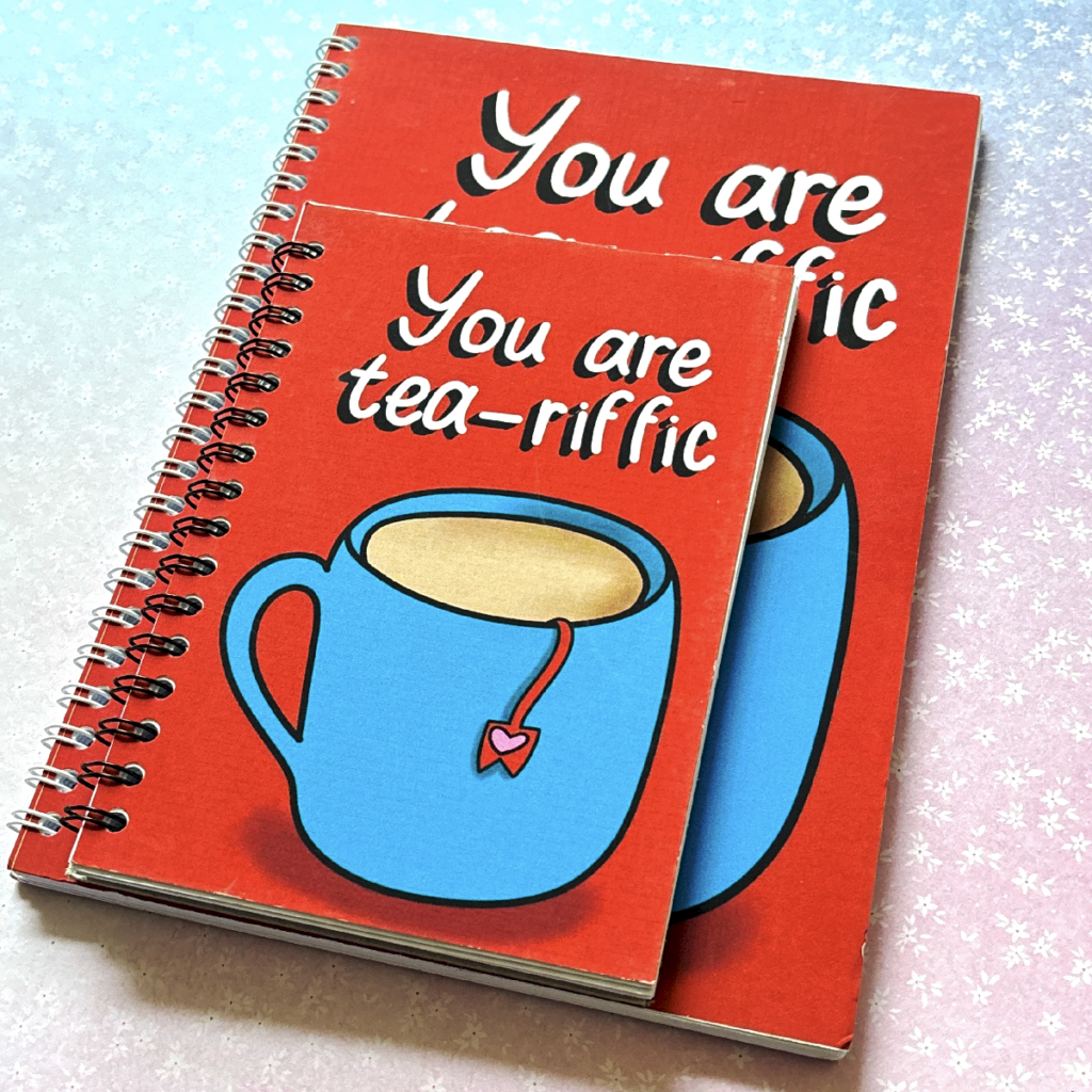 Two spiral bound notebooks lay on a pastel background. The notebooks have the same design, and the one on top is smaller than the bottom one. The notebook has a red background and a mug of tea illustration on it. Above is the phrase 'you are tea-riffic'.