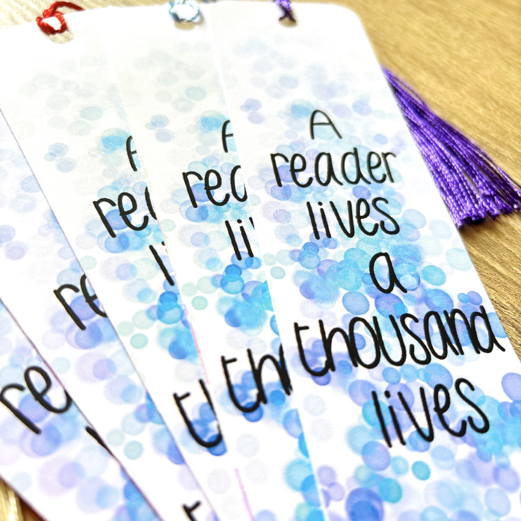 The Thousand Lives Bookmark Blue 3 features a blue and purple dot design, a purple tassel, and the quote "A reader lives a thousand lives." Four bookmarks are fanned out on a wooden surface.