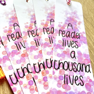 The Thousand Lives Bookmark Pink 3 features a pink and purple bokeh design with "A reader lives a thousand lives" in playful black lettering, beautifully displayed and fanned out on a wooden surface.