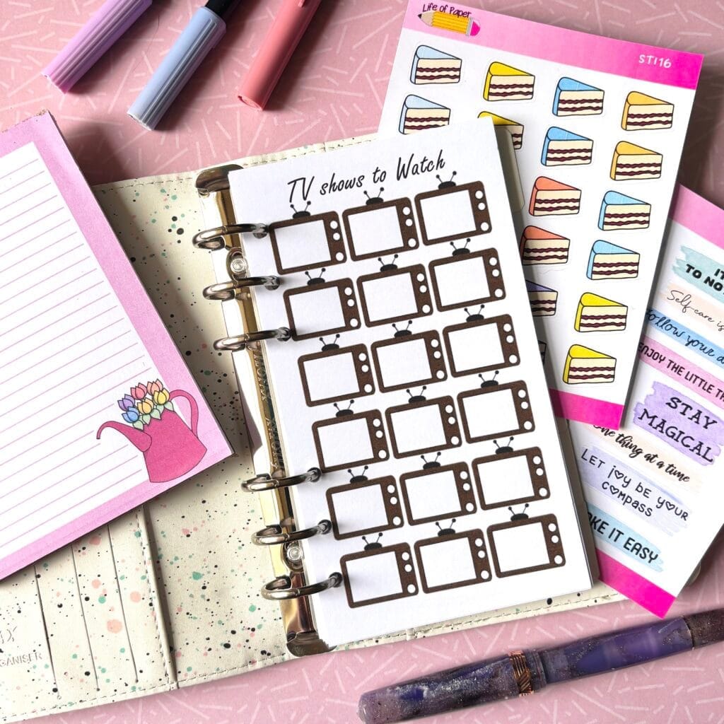 A personal ring planner open on a table. The top sheet shows a 'TV show tracker' insert. Around the planner are sticker sheets, a pink memo pad and some pens.