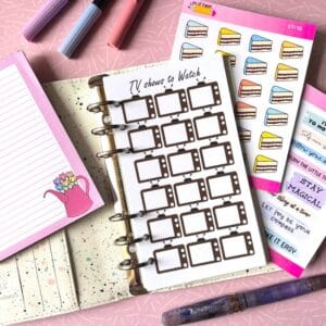A personal ring planner open on a table. The top sheet shows a 'TV show tracker' insert. Around the planner are sticker sheets, a pink memo pad and some pens.