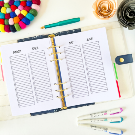 Year At A Glance Planner Insert A5 Ring 2 An A5 6-ring planner is open on a table. The top sheet has two months written at the top and below each month title is a table with a single column. The table has several rows and each row has a date on it, but no days. The picture has a cream background. Around the planner is a rainbow pompom coaster, roses made from card and highlighter pens.