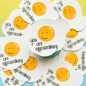 Stickers all with the same design are scattered all over the image with one raised slightly in the centre. A fried egg with a happy face drawn in the yoke and under is the text 'you are egg-traordinary'. The image has a blue to yellow hombre effect background.