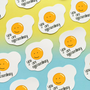 Stickers all with the same design are spaced evenly in diagonal rows going across the image. A fried egg with a happy face drawn in the yoke and under is the text 'you are egg-traordinary'. The image has a blue to yellow hombre effect background.