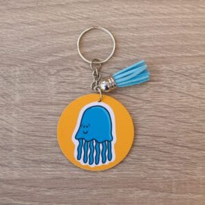 Jellyfish Keyring A blue jellyfish illustration on an orange background, The keyring is a circular shape and has a metal chain. There is a blue tassle on the keyring and it is lay on a light wooden surface.