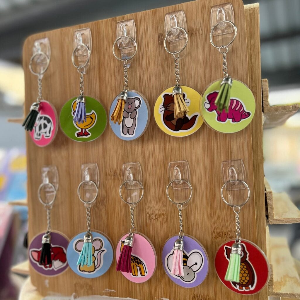 Ten keyrings hung up with plastic hooks on a wooden board. Each keyring has a coloured background and an animal illustration on it. Keyrings have a metal chain and a coloured tassle.