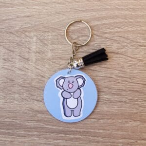 Koala Keyring Koala illustration on a pale blue background, The keyring is a circular shape and has a metal chain. There is a black tassle on the keyring and it is lay on a light wooden surface.