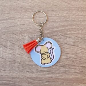 Keyring Field Mouse Mouse illustration on a pale blue background, The keyring is a circular shape and has a metal chain. There is a red tassle on the keyring and it is lay on a light wooden surface.