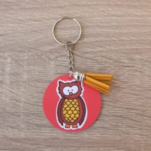 Owl Keyring Owl illustration on a red background, The keyring is a circular shape and has a metal chain. There is an orange tassle on the keyring and it is lay on a light wooden surface.
