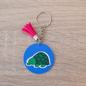Turtle Keyring Turtle illustration on a blue background, The keyring is a circular shape and has a metal chain. There is a red tassle on the keyring and it is lay on a light wooden surface.